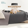 A light-colored dog is resting on a gray waterproof pet mat or sofa cover.