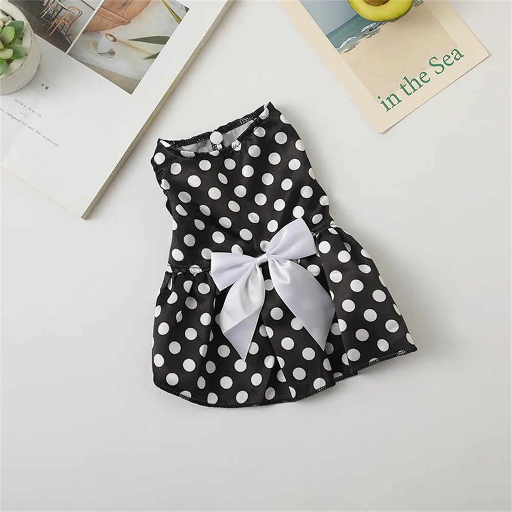 A black polka dot dog dress with a white bow, displayed on a light surface next to a book.