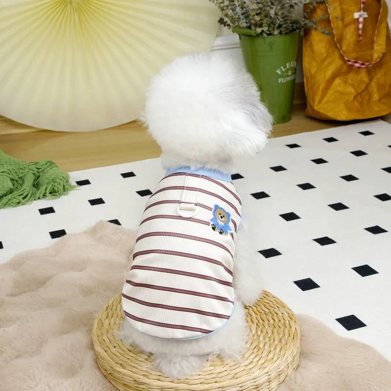 A white fluffy dog wearing a striped cooling vest with blue accents, sitting on a light beige surface.