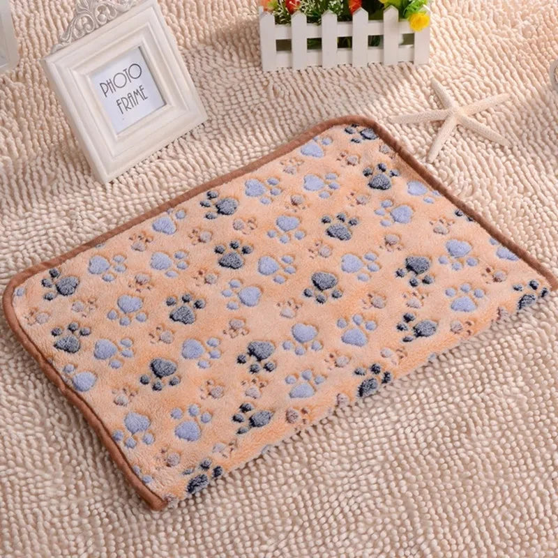 A square fleece pet bed mat cover with a light orange background and blue paw prints rests on a beige textured fabric.