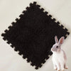 A small gray rabbit sits on a black, textured fleece pet warming mat.