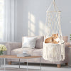A fluffy orange cat is resting inside a bohemian macrame hammock pet bed in a modern living room setting.