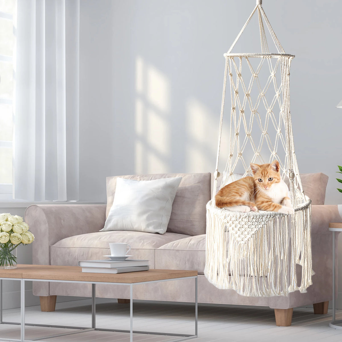 A fluffy orange cat is resting inside a bohemian macrame hammock pet bed in a modern living room setting.