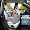 A small dog is sitting in a gray pet car seat inside a car, next to the driver's seat.
