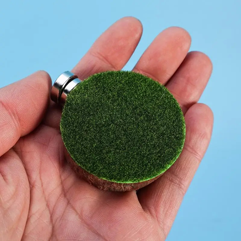 A hand holds a small, round piece of green mossy material attached to a metal ring.