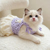 A fluffy white and light pink kitten wearing a purple floral print dress with thin straps.
