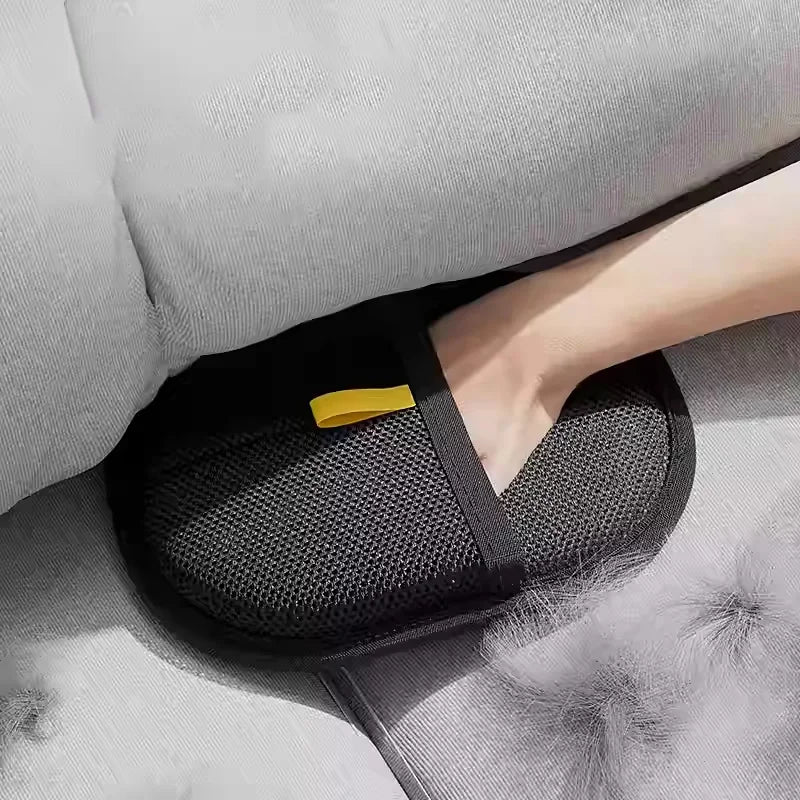 Una mano vistiendo guantes reutilizables para quitar pelo de mascotas negros con un acento amarillo, apoyada en un sofá gris.