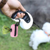 A hand holds a white reusable dog poop bag with a black handle and a pink tissue inside, against a blurred green background.