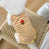 A white dog is wearing a yellow and pink striped floral print vest on a woven mat.