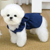 A fluffy white dog wearing a navy blue vintage corduroy pet dress with a bow and ruffle skirt.