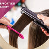 Une main utilise une peigne lissante rose en forme de V sur une mèche de cheveux brun foncé tenue par un fer plat noir.