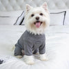 A white dog wearing a gray cozy pajama top lying on a white bed.