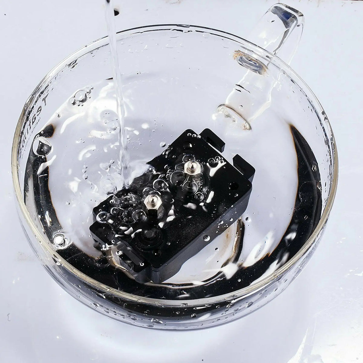 A black rechargeable dog training collar is being submerged in water inside a clear glass bowl.