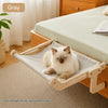 A fluffy light-colored cat is relaxing in a solid wood hanging cat hammock on a wooden bed.