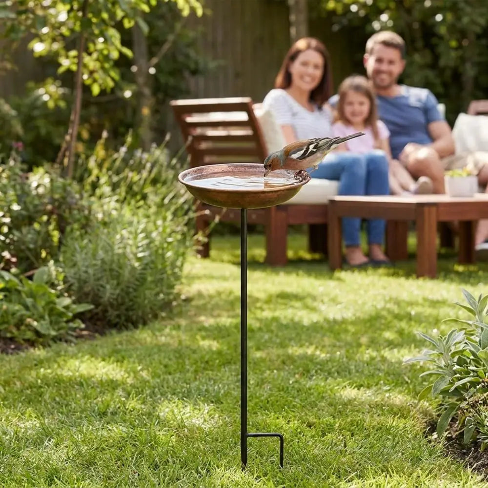 Une station d'alimentation pour oiseaux en métal décorative avec un bol, placée sur un support haut dans un jardin ensoleillé