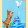 A fluffy orange cat interacts with a blue interactive toy featuring a feather wand and rolling balls.
