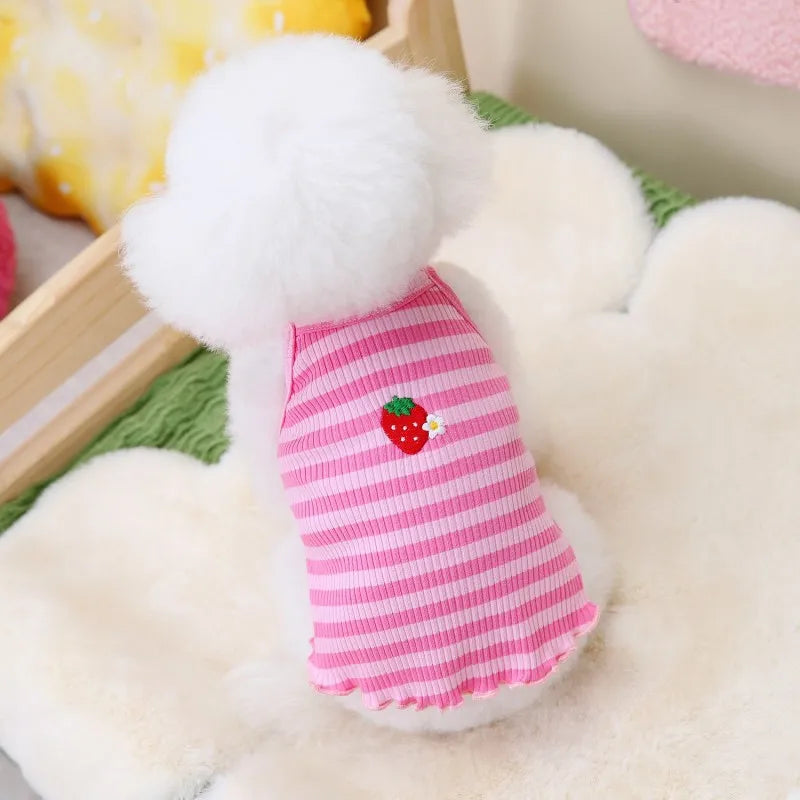 A white fluffy dog vest with a pink and white striped fabric and a red strawberry design is displayed on a white fur surface.