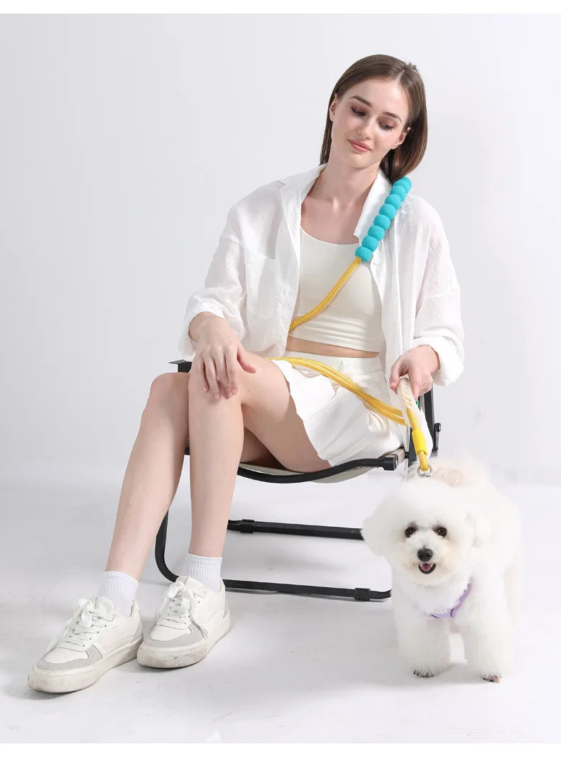 Una mujer está sentada en una silla con una correa y collar ajustable para perro sin manos, posando con un caniche blanco.
