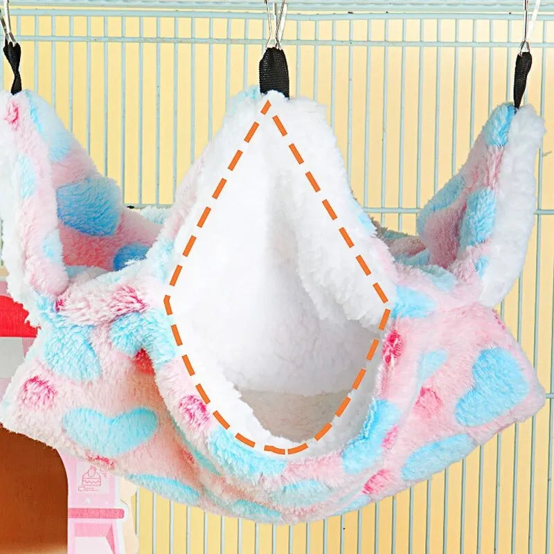 A pink and blue fluffy pet hammock with a white interior is hanging against a light yellow background.