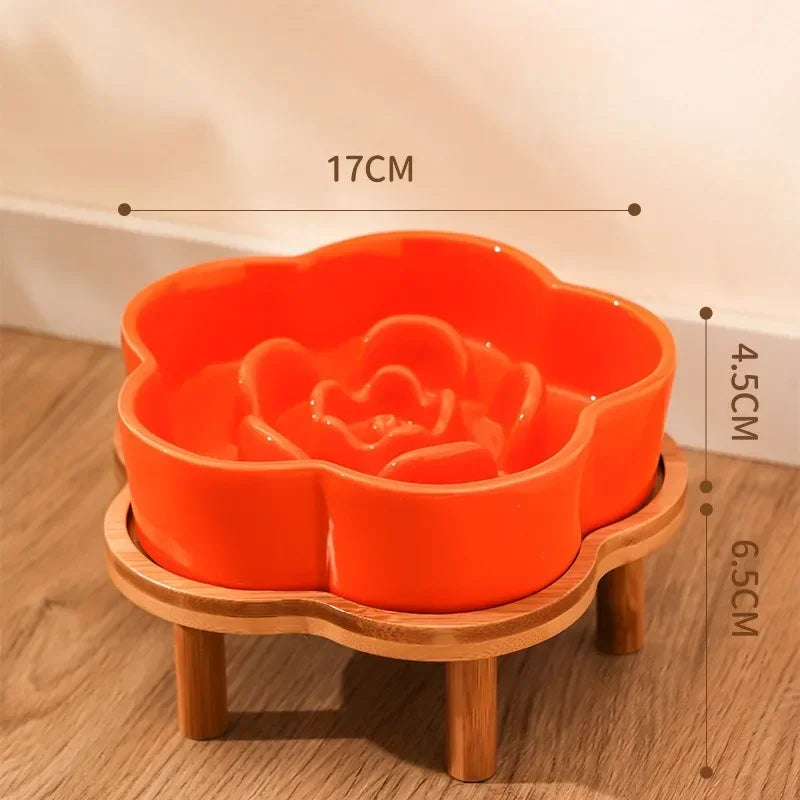 An orange ceramic slow feeder cat bowl rests on a natural wooden stand.