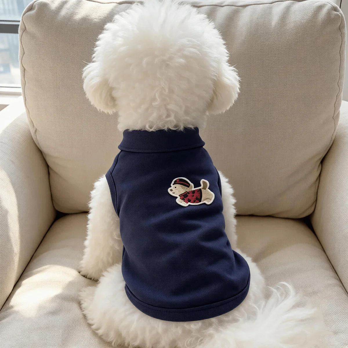 A white fluffy dog wearing a navy blue sleeveless vest with a cartoon embroidery on the back, sitting on a light-colored sofa