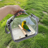 A hand holds a portable bird travel carrier with a transparent viewing panel and a wooden perch, set against a green grassy b
