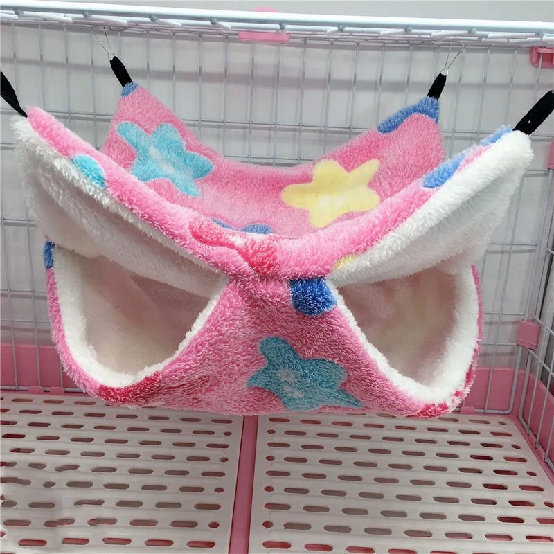 A pink and white fleece hammock bed with blue and yellow star patterns for a hamster is displayed in a wire cage.