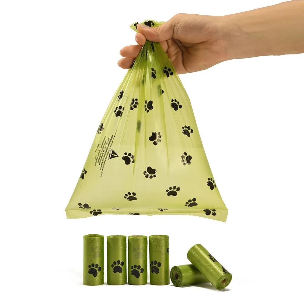 A hand is holding a clear biodegradable pet poop bag with black paw prints, next to several rolls of the same bags.