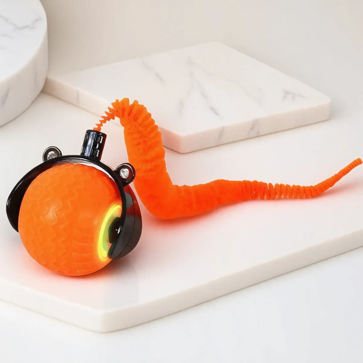An automatic rotating LED pet toy shaped like an orange worm resting on a white marble surface.