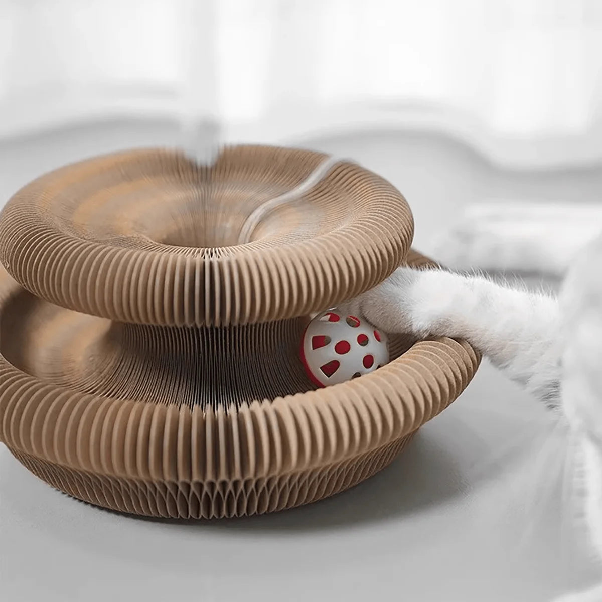 A close-up of a brown foldable accordion cat scratcher with a white catnip ball being held by a paw.