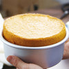 A hand holds a round, golden-yellow baked cake in a white aluminum pan.