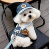 A white poodle wearing a blue and white plaid harness vest with a brown bear charm sitting on a black chair.