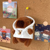 A brown capybara plush doll wearing a white hoodie with a small gold charm, displayed on a cork board.