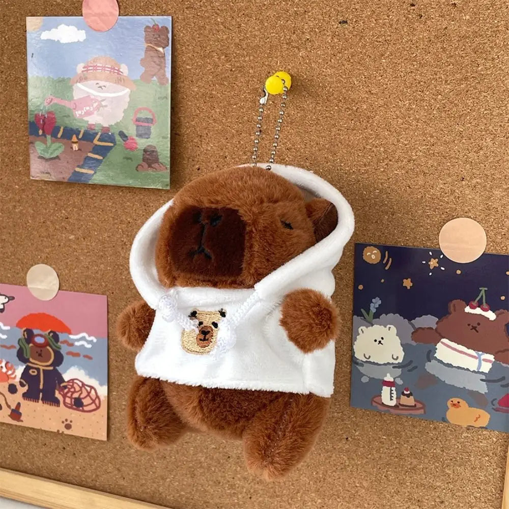 A brown capybara plush doll wearing a white hoodie with a small gold charm, displayed on a cork board.