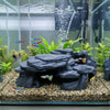 A realistic 3D printed fish tank rockery ornament featuring dark rocks and green plants with small colorful fish.