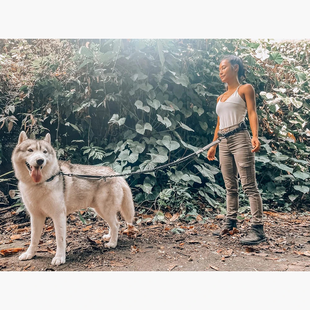 A woman is walking a fluffy white dog on a black leash in a wooded outdoor setting.