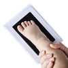 A handprint and footprint kit showing a baby's foot being pressed into a white frame by an adult's hand.