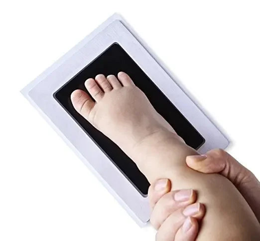 A handprint and footprint kit showing a baby's foot being pressed into a white frame by an adult's hand.
