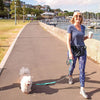 A woman walks a fluffy white dog on a hands-free reflective leash along a waterfront path.