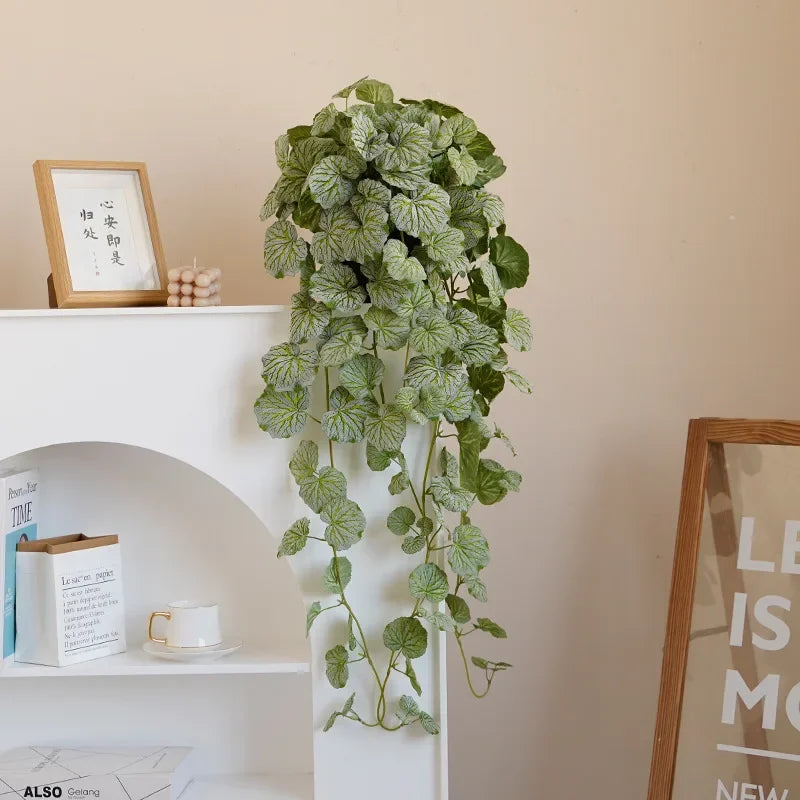 Künstliche Hängepflanzenranken mit grünen Blättern in einem weißen Topf vor einer hellen Wand.