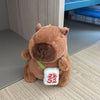 A brown capybara plush doll wearing a green strap and a white tag on its chest.