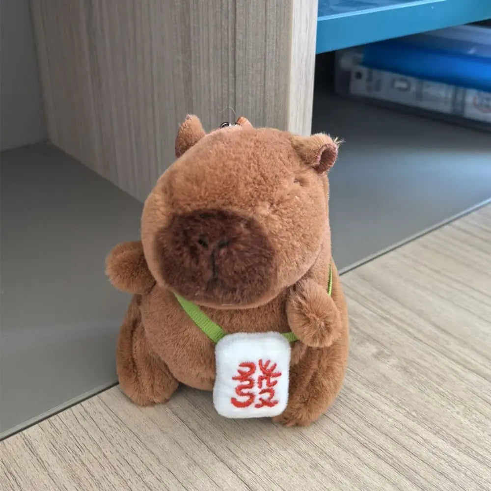 A brown capybara plush doll wearing a green strap and a white tag on its chest.