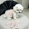 A fluffy white dog wearing a pink floral dress with a bow, sitting on a light gray surface.
