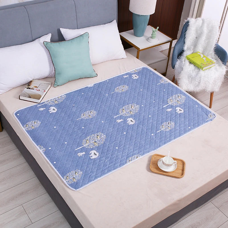 A blue patterned waterproof cartoon bed protector pad is placed on a light-colored table next to pillows and a white fluffy c