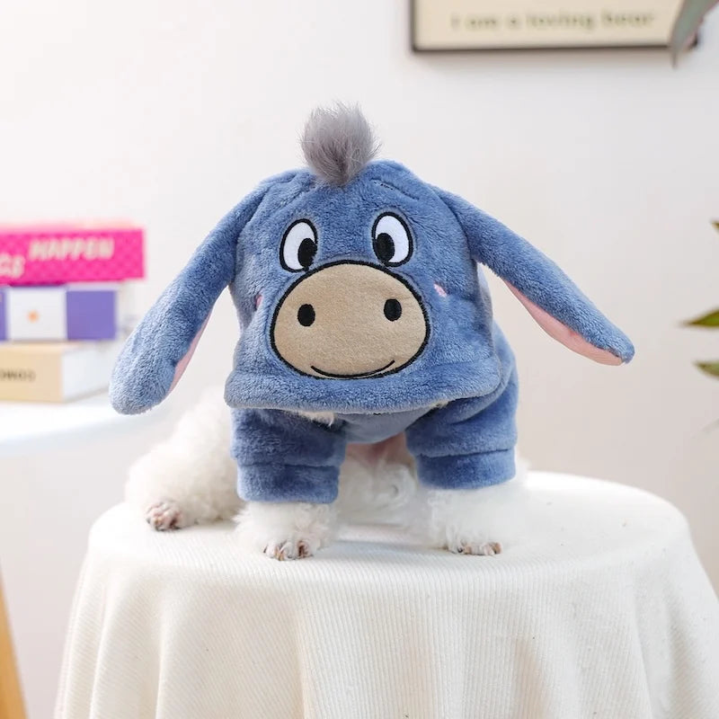A blue flannel hoodie shaped like a donkey is worn by a white dog sitting on a white table.