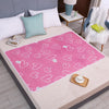 A pink heart-patterned waterproof bed protector pad is displayed on a light-colored table with pillows and a white fluffy bla
