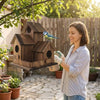 A woman is holding a rustic wooden bird house in her hands outdoors with greenery and a bird perched on the house.