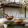 Two wooden bird feeders with hanging iron chains suspended from a tree branch over a water feature.