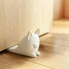 A white anti-slam door stopper wedge shaped like a cat rests on a wooden floor.