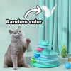 A gray cat sits next to an interactive toy tower with multiple turntables and a feather ball.